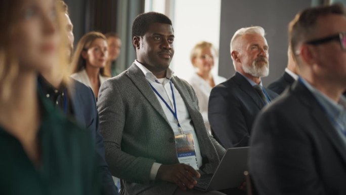 在商业会议上，一个黑人坐在拥挤的听众中。公司代表使用手提电脑聆听关于发展中市场的励志创业演讲。