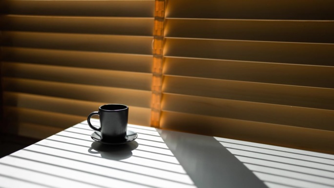 清晨日出时，明亮的阳光透过百叶窗照射进来，黑影出现在咖啡馆的黑色咖啡杯和咖啡桌上。