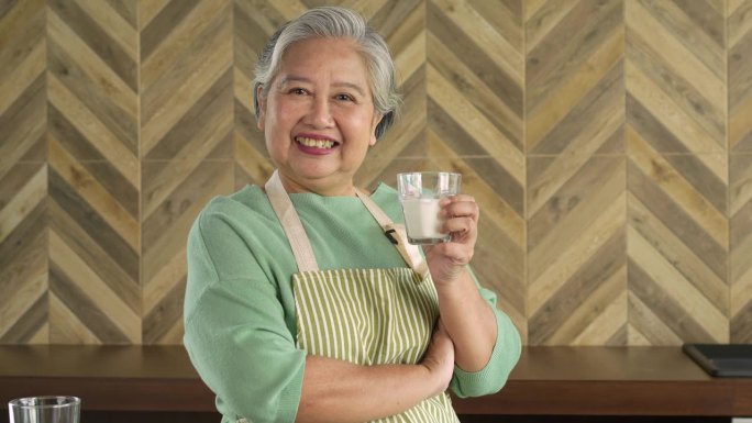 亚洲老年妇女早上在家里的厨房里喝着一杯牛奶，脸上挂着幸福的笑容。退休人员喝牛奶使骨骼强壮。预防骨质疏