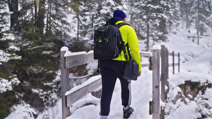 男背包客在白云石地区冰雪覆盖的森林里走在木桥上的后视图