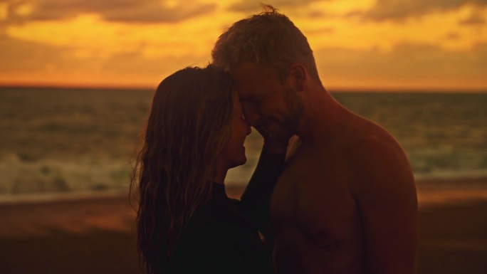 夕阳下，一对深情的冲浪夫妇在海滩上面对面。宁静，热爱冲浪夫妇拥抱面对宁静的海洋海滩在日落时分，享受夏