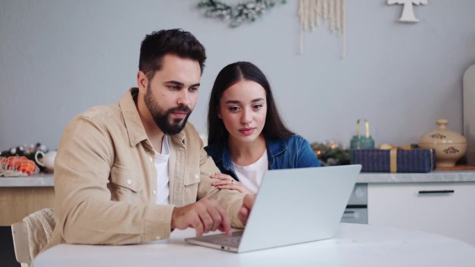 一对积极的西班牙夫妇一起在圣诞节装饰厨房网上购物，女人在笔记本电脑上选择礼物，在网上订购。快乐的多种
