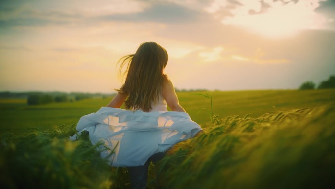 日落时分，棕色头发的兴奋女人在绿色的小麦地里奔跑