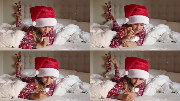 圣诞快乐，新年快乐。快乐的小女孩躺在床上装饰着家里的红帽子、绒球睡衣做想着给圣诞老人的信。孩子在给圣