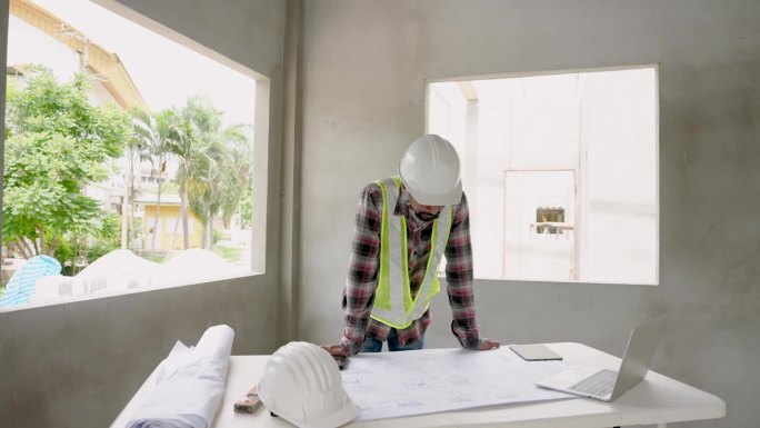 亚洲男性建筑师与建筑监理工人工程师雇员戴白色安全帽。查看结构图，并指出建筑物中需要固定的点。