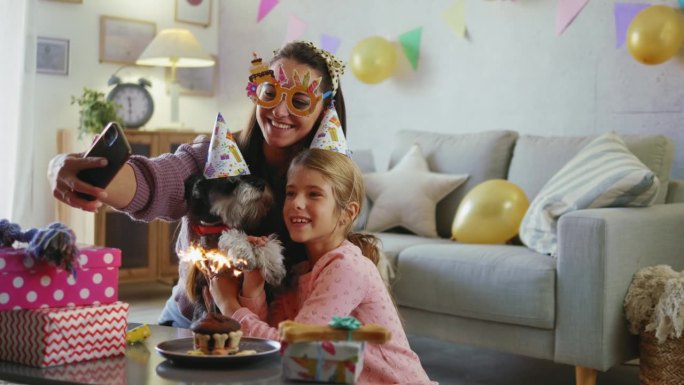 妈妈和女儿在家里为狗狗庆祝生日，戴上生日帽，吃上生日蛋糕，唱歌，自拍或拍摄社交媒体视频