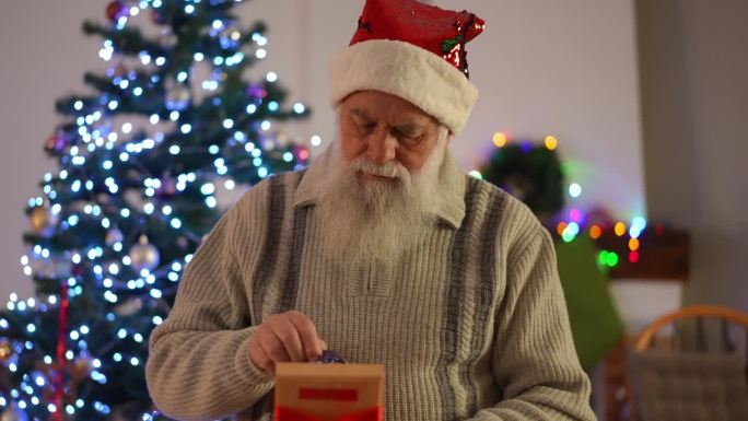 一位留着胡子、头戴圣诞帽的老人在圣诞树的背景上打开礼物。我们看到礼品盒里系着一条红丝带的男士领带。