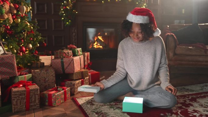 圣诞快乐。一位非裔美国妇女在圣诞树旁打开礼品盒。女孩在客厅用圣诞树和壁炉打开礼盒带着惊喜的脸，平安夜
