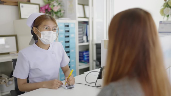 在诊所接待处，一名亚洲女士与一名护士谈论她的病情，并提供了她的个人资料和病史，护士将其记录在记事本上