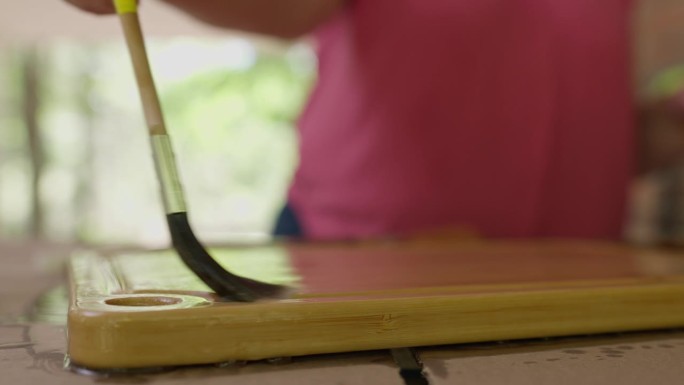 户外木艺:剪短女子双手浸渍，用矿物油涂刷木质砧板。模糊的背景，集中在刷和板。近距离