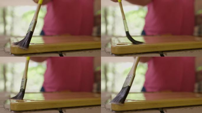 户外木艺:剪短女子双手浸渍，用矿物油涂刷木质砧板。模糊的背景，集中在刷和板。近距离