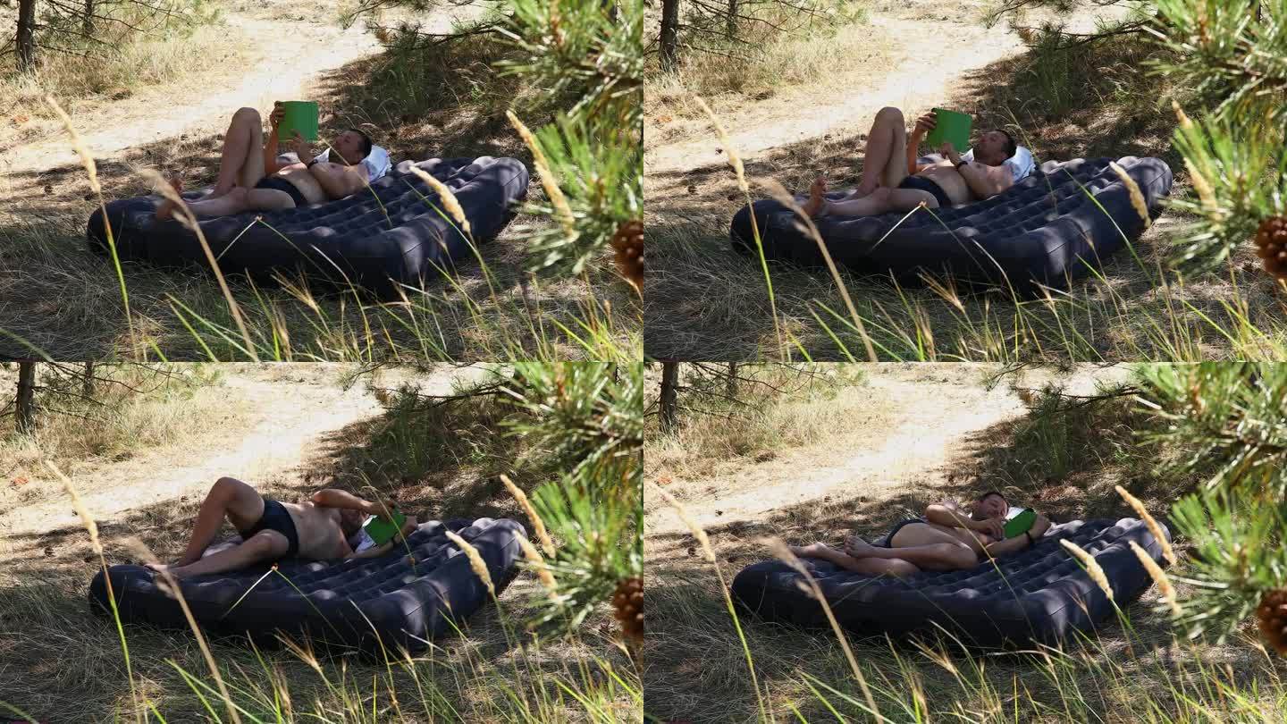 在一个阳光明媚的夏日，一个男人躺在树林里的充气床垫上看书。