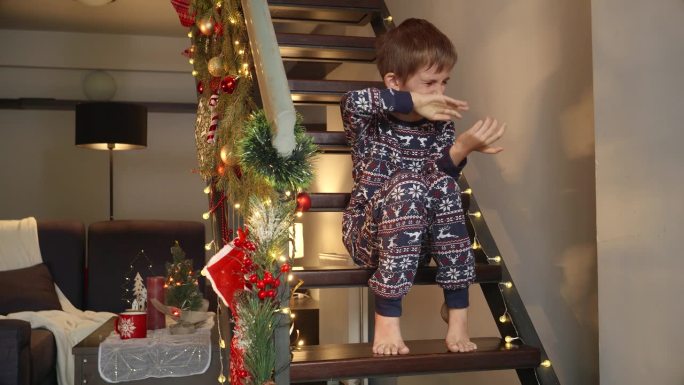 穿着睡衣的小男孩在圣诞夜独自坐在木楼梯上哭泣。冬季假期和新年的悲伤、抑郁、孤独和负面情绪