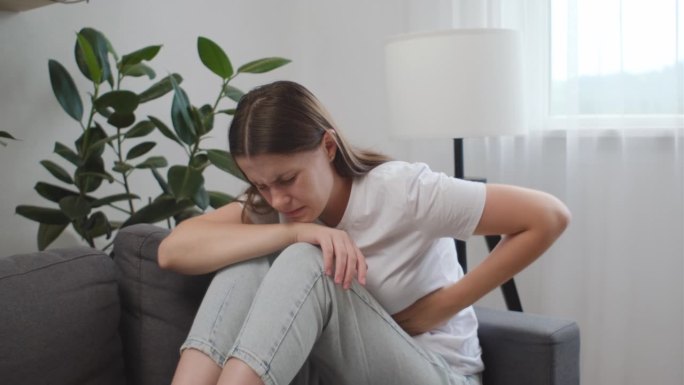 患有胃痛的不健康的年轻白人妇女坐在沙发上。心烦意乱的黑发女孩抱着肚子，感觉腹部月经疼痛或肠道和消化问