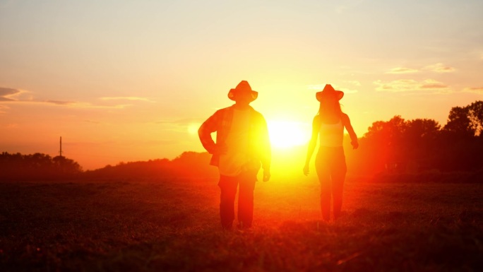 两个戴草帽的农民的剪影，在红色的夕阳下走过耕地。夫妻俩经营家族农业企业。合作伙伴散步和检查他们的农村