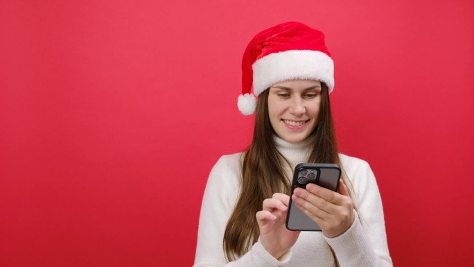 25岁的快乐年轻女子穿着白色毛衣，手拿圣诞帽，手持手机，在淡红色的工作室背景下孤独地摆姿势。2024