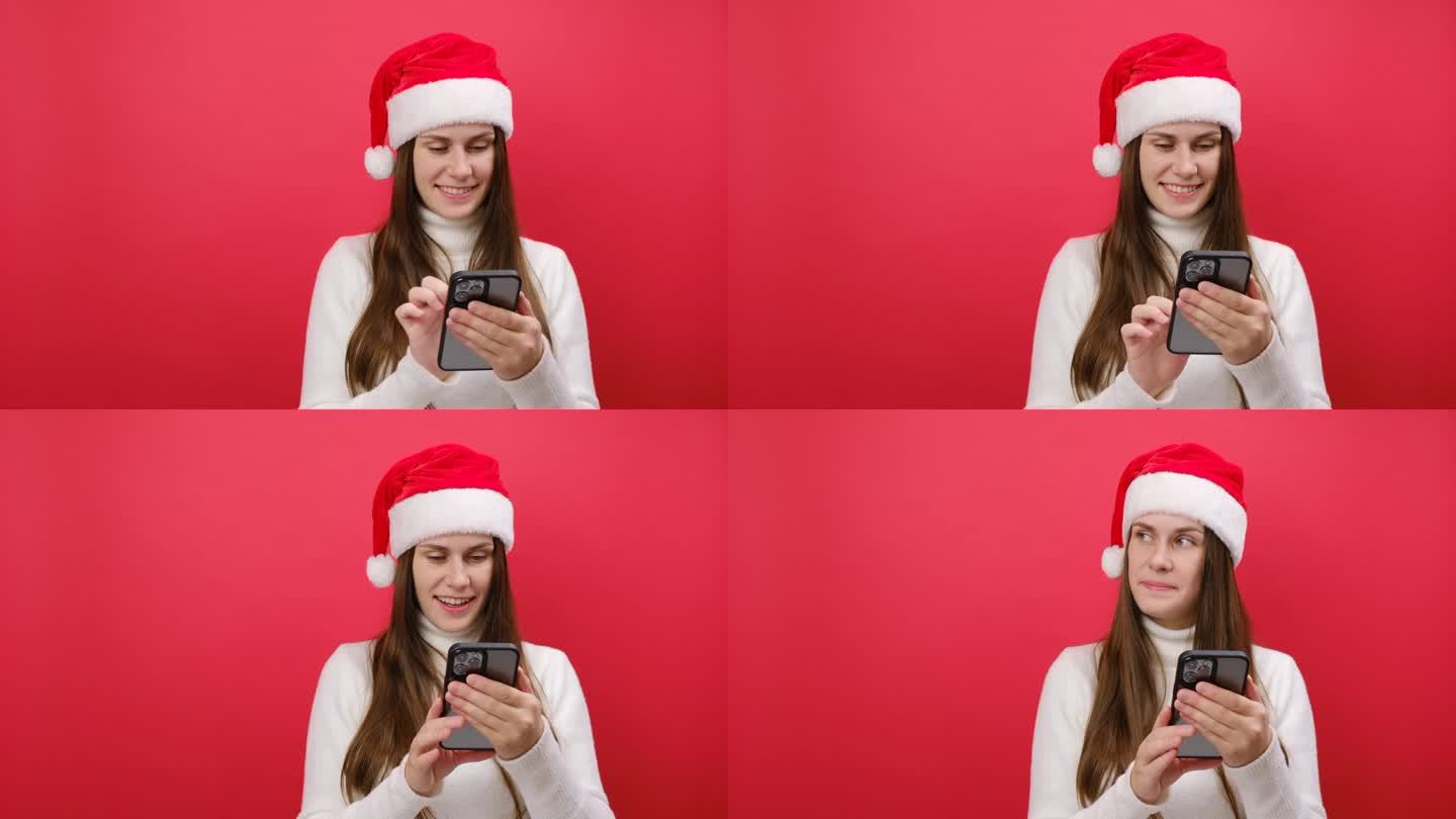 25岁的快乐年轻女子穿着白色毛衣，手拿圣诞帽，手持手机，在淡红色的工作室背景下孤独地摆姿势。2024