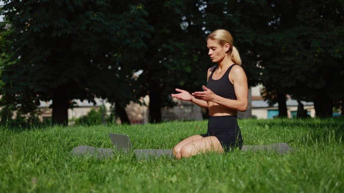 肌肉发达的白人女性坐在公园里的黑色瑜伽垫上，用无线笔记本电脑录制视频，讲述训练规则。爱运动的女性在健