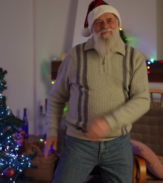 垂直视频中，一位老人穿着牛仔裤、毛衣和圣诞帽，在一棵圣诞树旁跳舞。