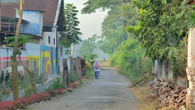 一位身穿制服、头戴头巾的印尼女学生走在一条破旧的乡间小路上，远处是一片泛黄的稻田