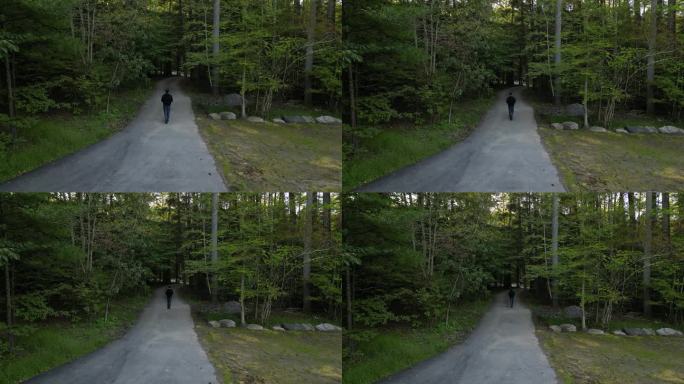 航拍画面显示，一名男子背着包裹，双手被占着，在乡间郊区一条被树木环绕的小路上走着(4k无人机从上方拍