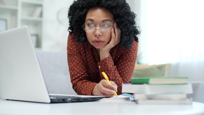 疲惫的非裔美国女学生坐在家里的客厅里用笔记本电脑学习。
