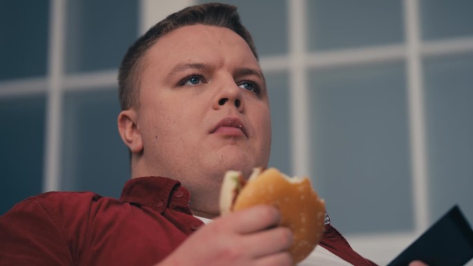 超重的年轻人一边嚼汉堡一边看电视，吃太多垃圾食品