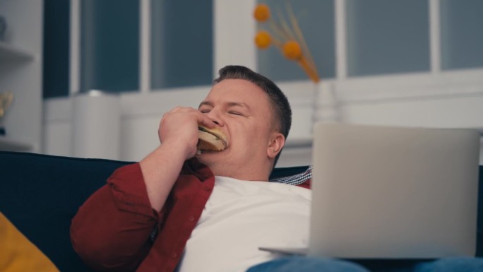 不整洁的超重男子吃着油腻的垃圾食品，坐在沙发上用笔记本电脑