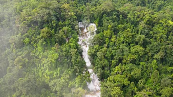 从空中俯瞰琅勃拉邦的热带森林树木环绕的光寺瀑布。后拉镜头与倾斜，通过云层揭示森林景观