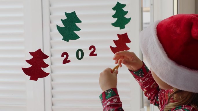 小女孩戴着红色的圣诞老人帽子装饰冬季度假屋，为新年派对手工纸花环数字2024圣诞树。孩子们自己动手做