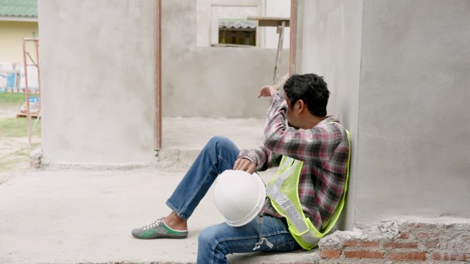 印度男子经理工头工人工程师住房工程建筑工程靠墙坐着，膝盖弯曲，摘下白色头盔，擦去汗水。在施工区内
