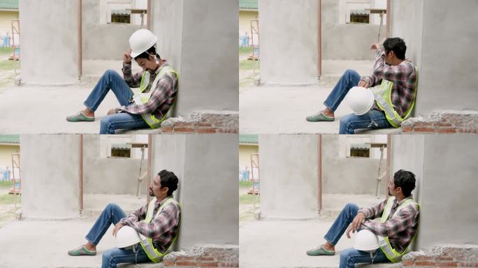 印度男子经理工头工人工程师住房工程建筑工程靠墙坐着，膝盖弯曲，摘下白色头盔，擦去汗水。在施工区内