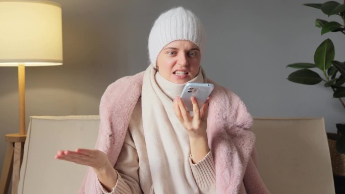 愤怒的女人手里拿着手机，穿着保暖的外套和帽子，与中央供暖系统争吵，手里拿着智能手机，公寓里的暖气出现