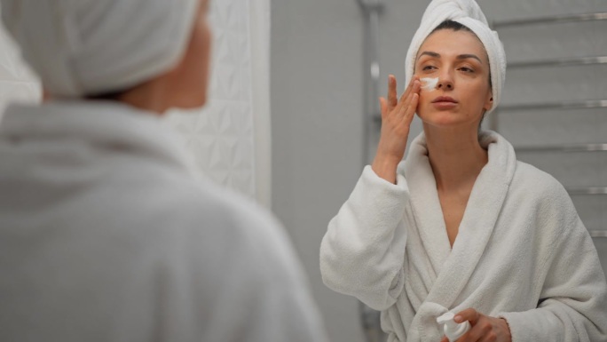 女士使用保湿泡沫洗脸和微笑，早上的护肤程序。年轻的女学生头上缠着毛巾，在水疗沙龙照镜子。准备化妆。