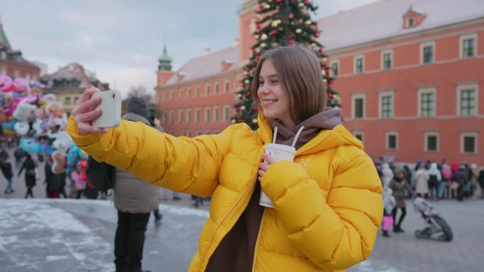 穿着黄色外套的少女在市镇广场的圣诞树背景下自拍