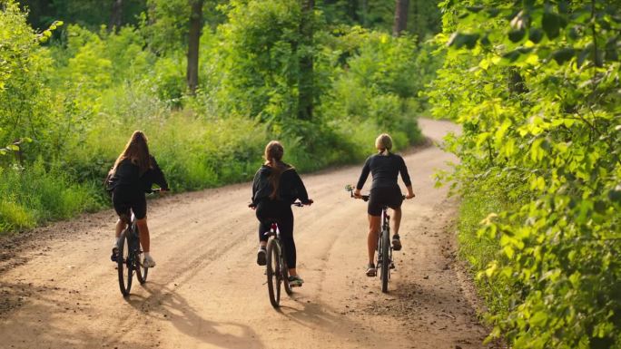 三个年轻女子骑自行车在地面道路上的森林，后景，慢镜头在大自然