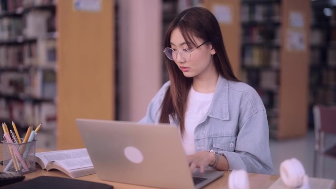 勤奋的亚洲女学生在笔记本电脑上输入她的研究报告，输入从大学图书馆的书中收集的信息。展示学术承诺，利用