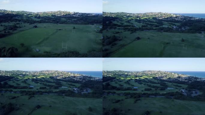 无人机飞过橄榄球场，俯瞰绿色风景的Bluff高尔夫球场，背景是道路分割和海洋