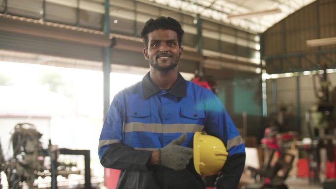 中年黑人工程师手持安全帽在工厂内看镜头，背景是移动的机械臂