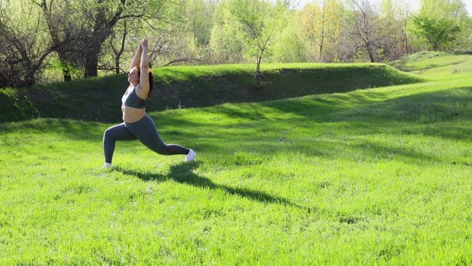 阳光明媚的一天，在公园的绿色草坪上，穿着健身服的年轻超重女子用脚弓步，举起手臂，慢动作。健康的生活方