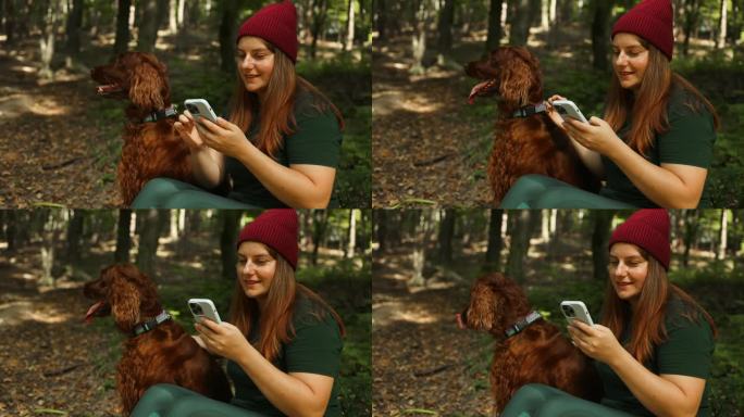 一个快乐的女人，穿着时尚的运动服，背着背包，坐在一棵树上，在森林里拿着手机。一只美丽的红色爱尔兰塞特