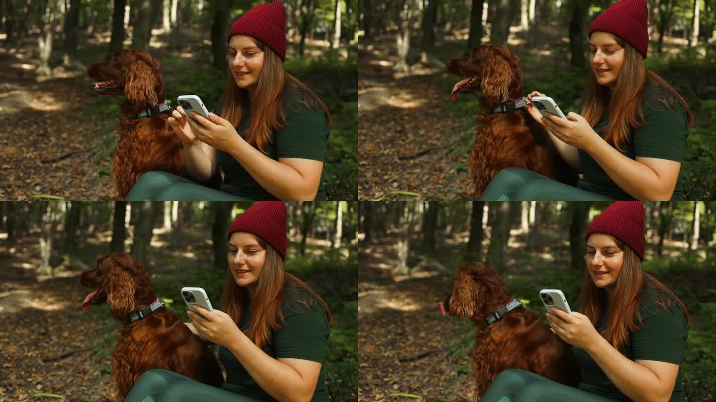 一个快乐的女人，穿着时尚的运动服，背着背包，坐在一棵树上，在森林里拿着手机。一只美丽的红色爱尔兰塞特