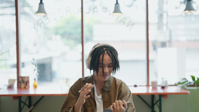 亚洲年轻自由职业者戴着白色耳机，在一个阳光明媚的日子里在咖啡馆里使用平板电脑