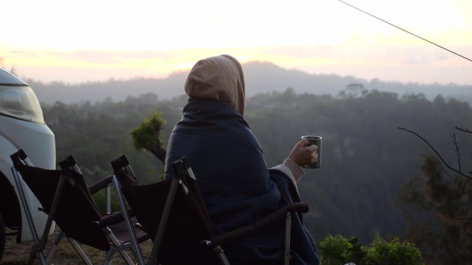 一个悲伤的女人端着一杯咖啡坐在房车旁，俯瞰着黎明的群山和森林。房车旅行中孤独生活的特写，享受野生动物