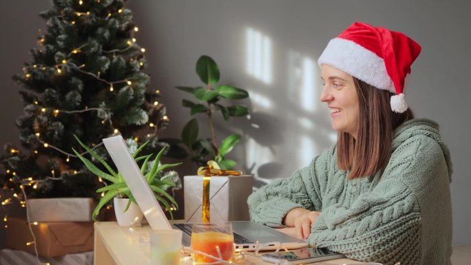 友好的高兴的女人穿着绿色针织衫和圣诞老人帽在圣诞装饰的房间里视频通话，挥手问候，微笑愉快地与朋友谈论