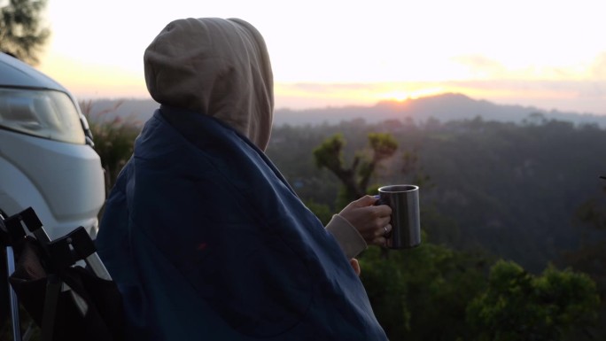 一个女人端着一杯咖啡坐在房车旁，在黎明的雾中俯瞰群山。房车旅行中平静生活的特写，享受野生动物景观。乘