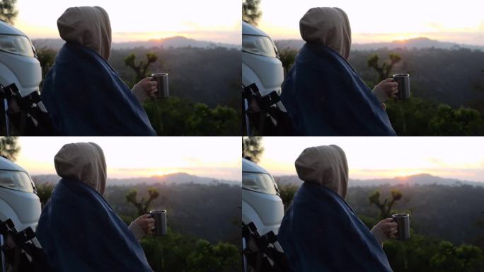 一个女人端着一杯咖啡坐在房车旁，在黎明的雾中俯瞰群山。房车旅行中平静生活的特写，享受野生动物景观。乘