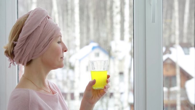 一个戴头巾的女人在窗外冬日的背景下喝着黄色的可溶性维生素饮料。