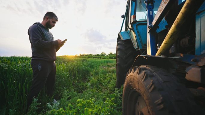 田地里的人拿着电话站在拖拉机旁敲打车轮，检查机器状况以诊断设备的可用性。经营一家卖农产品的小公司。