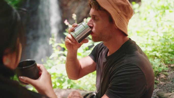 放松的游客夫妇用保温瓶喝茶，在瀑布边聊天